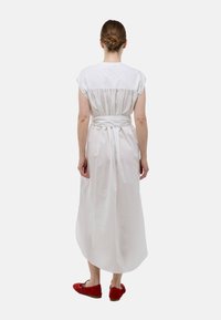 Robe en coton blanche avec des manches courtes, dos froncé et ceinture à la taille. Portée avec des chaussures rouges, elle présente un design fluide de longueur cheville.