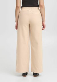 Pantaloni beige a gamba ampia con vita elastica, texture liscia e dettaglio della tasca posteriore. Indossati con calzature nere.