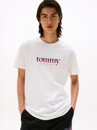 T-shirt en coton blanc avec le logo "tommy JEANS" en rouge et bleu. Manches courtes, col ras du cou, coupe décontractée. Le modèle se tient avec les mains dans les poches.
