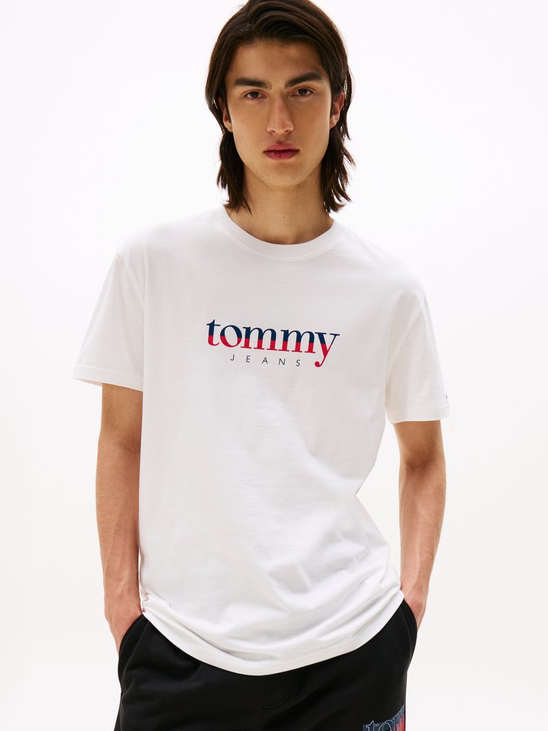 T-shirt en coton blanc avec le logo "tommy JEANS" en rouge et bleu. Manches courtes, col ras du cou, coupe décontractée. Le modèle se tient avec les mains dans les poches.