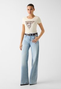 Kremowy dopasowany t-shirt z trójkątnym logo "GUESS" i cekinami, zestawiony z jasnoniebieskimi jeansami o szerokich nogawkach i ciemnobrązowym paskiem.