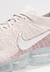 Nike-sneakers met een gestructureerd lichtbeige gebreid bovenwerk, een zilveren Swoosh-logo en een transparante zool met luchtkussen, voorzien van een roze verlopend accent.