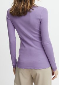 Top de punto largo de color púrpura con dobladillo en forma de onda y detalles de encaje en los puños, combinado con pantalones de color claro. Textura de tela suave.