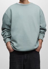 Sudadera verde menta con un corte holgado, cuello redondo y puños acanalados, combinada con jeans oscuros. Textura suave y diseño casual.