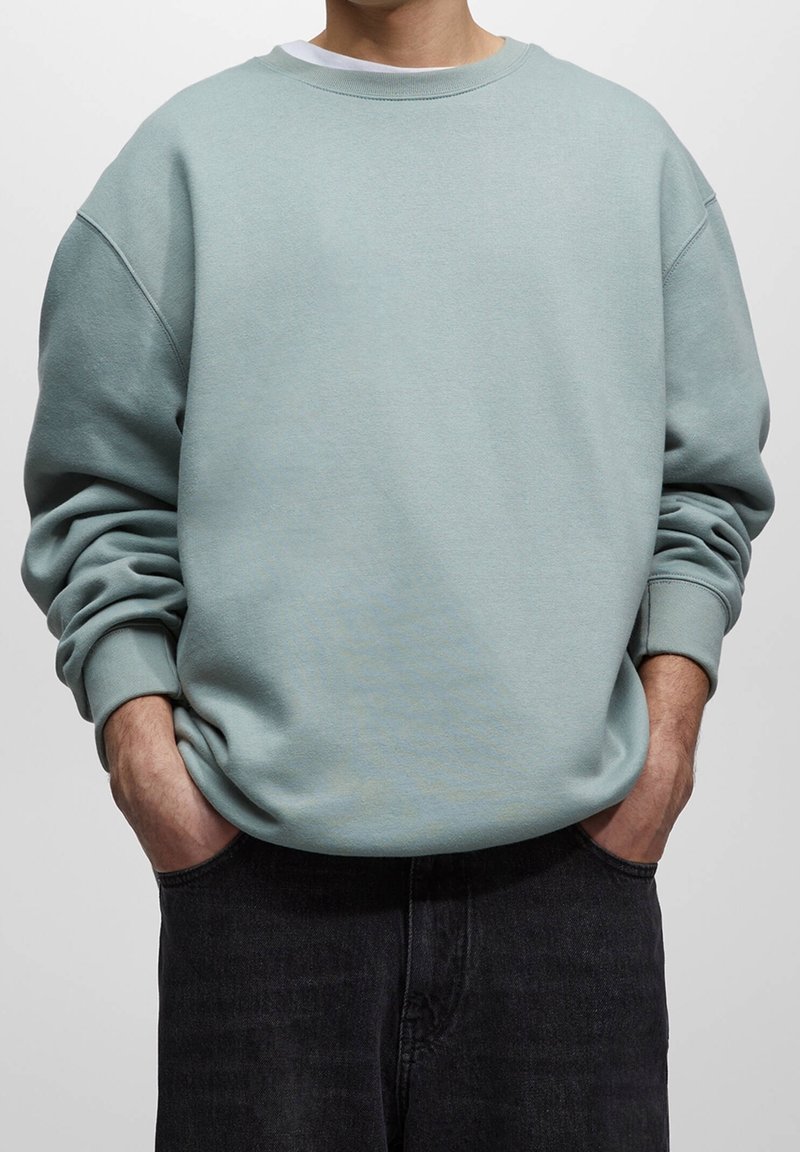Sudadera verde menta con un corte holgado, cuello redondo y puños acanalados, combinada con jeans oscuros. Textura suave y diseño casual.