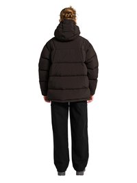 Osoba związaną fryzurą, ubrana w czarną pikowaną kurtkę z kapturem i luźne czarne spodnie, stojąca tyłem do przodu.