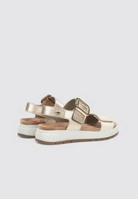 Sandalias de mujer con parte superior metálica dorada, correa ajustable y suela blanca texturada. Presenta una plantilla plana y suave y un detalle de hebilla.