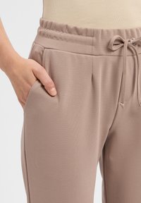 Jasnobrązowe spodnie dresowe z elastycznym pasem, detalem sznurkowym i bocznymi kieszeniami, wykonane z gładkiego, elastycznego materiału.