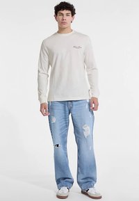 Kremowa koszula z długim rękawem z bawełny z małym logo, zestawiona z jasnoniebieskimi, przetartymi jeansami z przetarciami, dopełniona białymi trampkami.