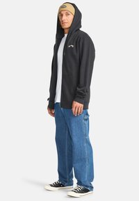 Schwarze Zip-Hoodie mit einer Vorderentasche und weißem Logo, kombiniert mit hellblauen Denim-Cargohosen und schwarzen Sneakers mit weißen Schnürsenkeln.