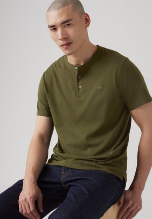 Olivgrünes Kurzarm-Henley-Shirt mit einem runden Ausschnitt, einer Zwei-Knopf-Leiste und einem dezenten Logo-Detail auf der Brust. Kombiniert mit dunklen Jeans.