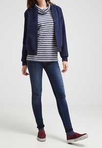 Marinblå bomberjacka över en grå och vit randig cowl neck-topp, kombinerad med blå skinny jeans och röda slip-on skor.