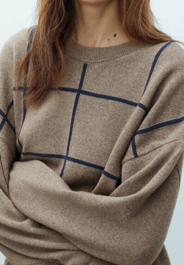 Femme portant un pull beige avec des lignes de quadrillage bleues, les bras croisés et de longs cheveux bruns tombant sur ses épaules.