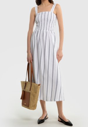Mujer con vestido midi blanco sin mangas a rayas, sosteniendo un bolso tote tejido color tostado y con zapatos planos slingback negros sobre fondo gris.