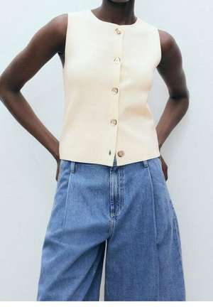 Haut beige sans manches à boutons, assorti à un jean en denim bleu taille haute à jambes larges, porté par une personne avec les mains sur les hanches.