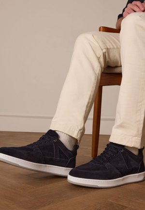 Baskets en suède bleu marine avec semelle en caoutchouc blanc, présentant des accents géométriques et six œillets, portées avec un pantalon ajusté de couleur claire.