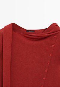 Red knit sweater with a wide neckline, diagonal row of matching red buttons, and a long fabric belt beside the collar.