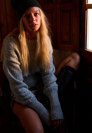 Strikket grå og blå sweater med lange ærmer, parret med en mørk hue. Model siddende ved et trævindue, med en struktureret væg i baggrunden.