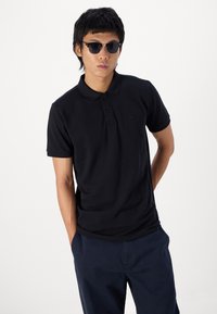 Black polo shirt made of smooth fabric, featuring a collar and three-button placket. Worn with dark blue trousers and black sunglasses.