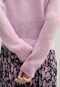Rosa gestrickter Pullover mit einer lockeren, luftigen Textur und gerippten Bündchen. Getragen über einem gemusterten, blümchenfarbenen, plissierten Rock, der die Schichtung von Stilen betont.