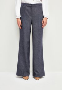 Bredbenta byxor i en marin herringbone-mönster, tillverkade av texturerad tyg. Har ett slätt midjeband och kombineras med spetsiga tanfärgade skor.