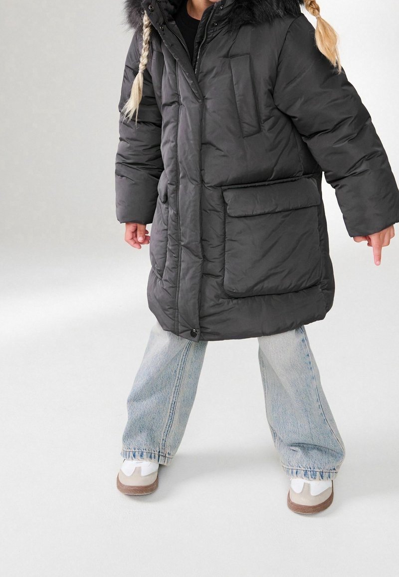 Enfant portant un long manteau matelassé noir avec de grandes poches, un jean bleu clair, des baskets blanches et deux tresses blondes sur un fond uni.