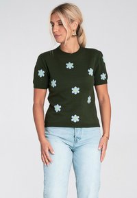 Kobieta w ciemnozielonym swetrze z krótkim rękawem, w jasnoniebieskie kwiaty, zestawiona z jasnoniebieskimi dżinsami, patrząca z boku na tle białym.