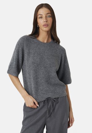 Vrouw met lang bruin haar, gekleed in een losse grijze kortemouwen trui en grijze krijtstreep broek met trekkoord, staand voor een effen achtergrond.