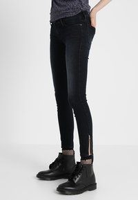 Jeans negros ajustados con detalles de cremallera en el tobillo, hechos de mezclilla, que presentan una textura suave y un diseño ajustado. Combinados con botas negras.