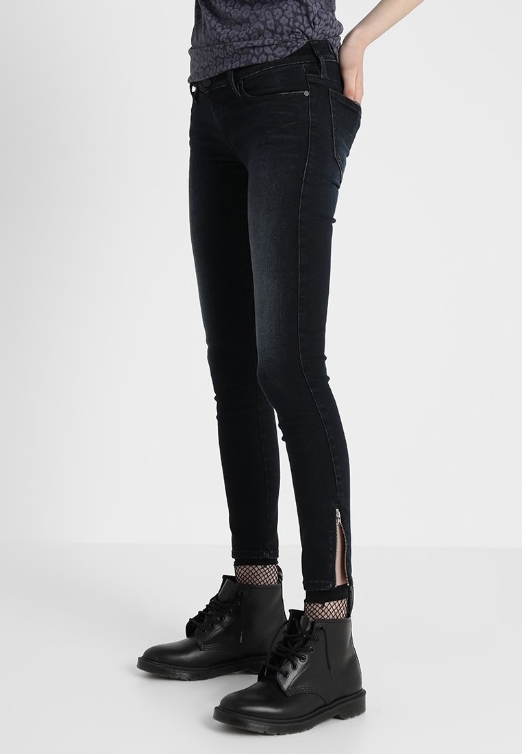 Jeans negros ajustados con detalles de cremallera en el tobillo, hechos de mezclilla, que presentan una textura suave y un diseño ajustado. Combinados con botas negras.