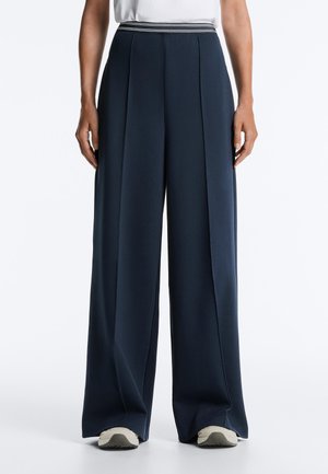 Marineblaue Hose mit weitem Bein und vorderen Bügelfalten, getragen mit weißen Turnschuhen und einem in die Hose gesteckten weißen Hemd, von der Taille bis zu den Füßen gezeigt.