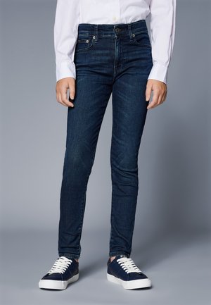 Osoba nosící tmavě modré džíny s úzkým střihem, bílou košili na knoflíky a tmavě modré tenisky s bílými tkaničkami na šedém pozadí.