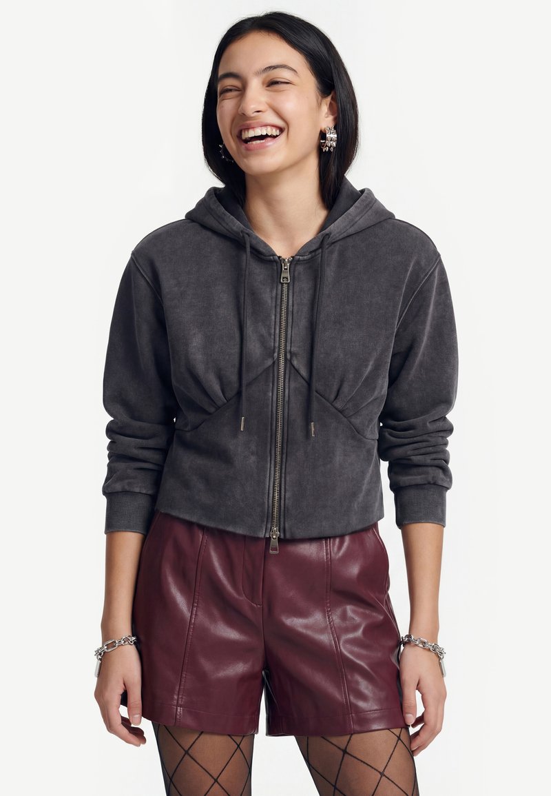 Jeune femme souriante portant un sweat à capuche zippé gris foncé, un short en cuir bordeaux, des collants résille noirs, des boucles d'oreilles anneaux argentées et des bracelets.