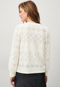 Blouse blanche en dentelle, ornée d'un motif en diamants avec des détails ajourés et des manches longues. Texture lisse avec une coupe décontractée.