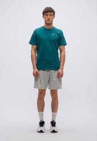 Τυρκουάζ βαμβακερό μπλουζάκι με στρογγυλή λαιμόκοψη, συνδυασμένο με ανοιχτό γκρι σορτς. Φορεμένο με ασπρόμαυρα αθλητικά παπούτσια, με λευκές κάλτσες ορατές.