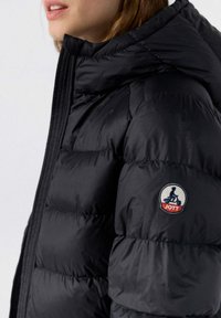 Kvinde iført en sort pufferjakke med høj krave og et JOTT-logo patch, der viser en person på en scooter på den øverste ærme.