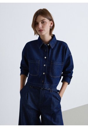 Femme portant une veste en jean bleu foncé avec poches avant et un pantalon large taille haute assorti, mains dans les poches, fond neutre.