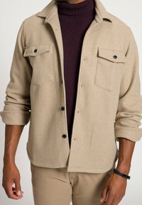 Beżowa kurtka polarowa z dwoma kieszeniami na piersiach, czarnymi guzikami i podwiniętymi rękawami, noszona na ciemnym bordowym golfie. Miękka, teksturowana tkanina.