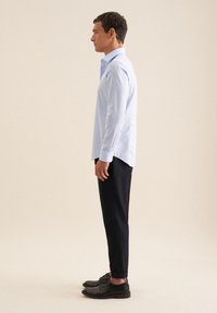 Camicia azzurra a maniche lunghe con colletto a punta, abbinata a pantaloni scuri e scarpe nere. La camicia presenta delle fessure laterali all'orlo.