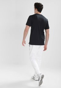 Camiseta negra de manga corta, textura suave, combinada con pantalones de chándal blancos, mostrando un ajuste relajado y un detalle de bolsillo lateral, vista desde atrás.