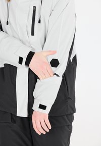 Grau- und schwarze wasserdichte Jacke mit einer Reißverschlusstasche, verstellbaren Manschetten und hexagonalem Logo-Detail, mit glatter Textur und lockerer Passform.