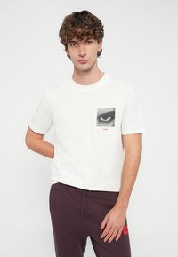 Biały bawełniany t-shirt z kwadratową czarno-białą grafiką oka oraz czerwonym napisem "HUGO". Model nosi ciemnofioletowe spodnie dresowe.