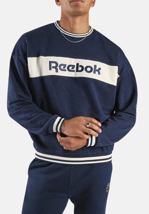 Hombre vistiendo sudadera azul marino de Reebok y pantalones a juego con puños y cintura de rayas blancas, de pie con las manos entrelazadas.
