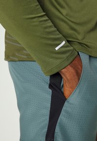 Hand in der Tasche kurzer, hellblauer Shorts mit schwarzem Seitenpanel, trägt ein langärmliges, olivgrünes Shirt mit kleinem weißen Logo am Ärmel.
