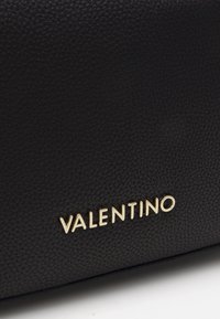 Svart texturerad läderväska med en guld "VALENTINO"-logotyp. Närbild som framhäver material och brandingdetalj.