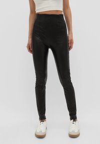 Leggings de imitación cuero negro de cintura alta con un diseño ajustado, textura suave y detalles acanalados, combinados con zapatillas blancas.
