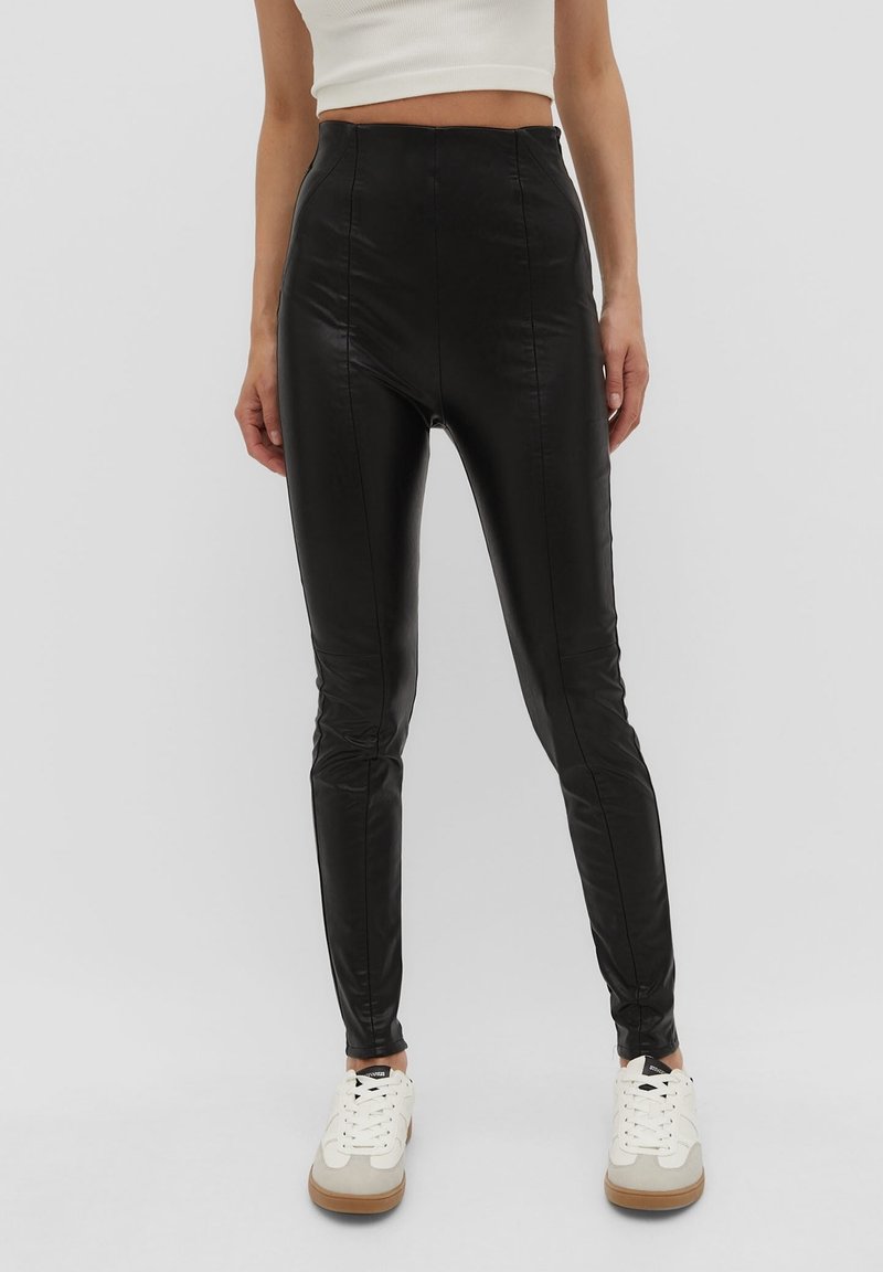 Leggings de imitación cuero negro de cintura alta con un diseño ajustado, textura suave y detalles acanalados, combinados con zapatillas blancas.