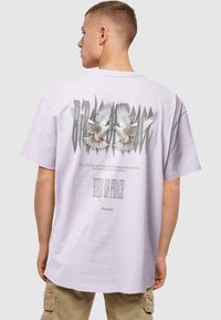 Lilla oversized t-shirt med et grafik af to hvide duer og stiliseret tekst "FUGL AF FRED" på bagsiden. Korte ærmer, lavet af bomuldsmateriale.