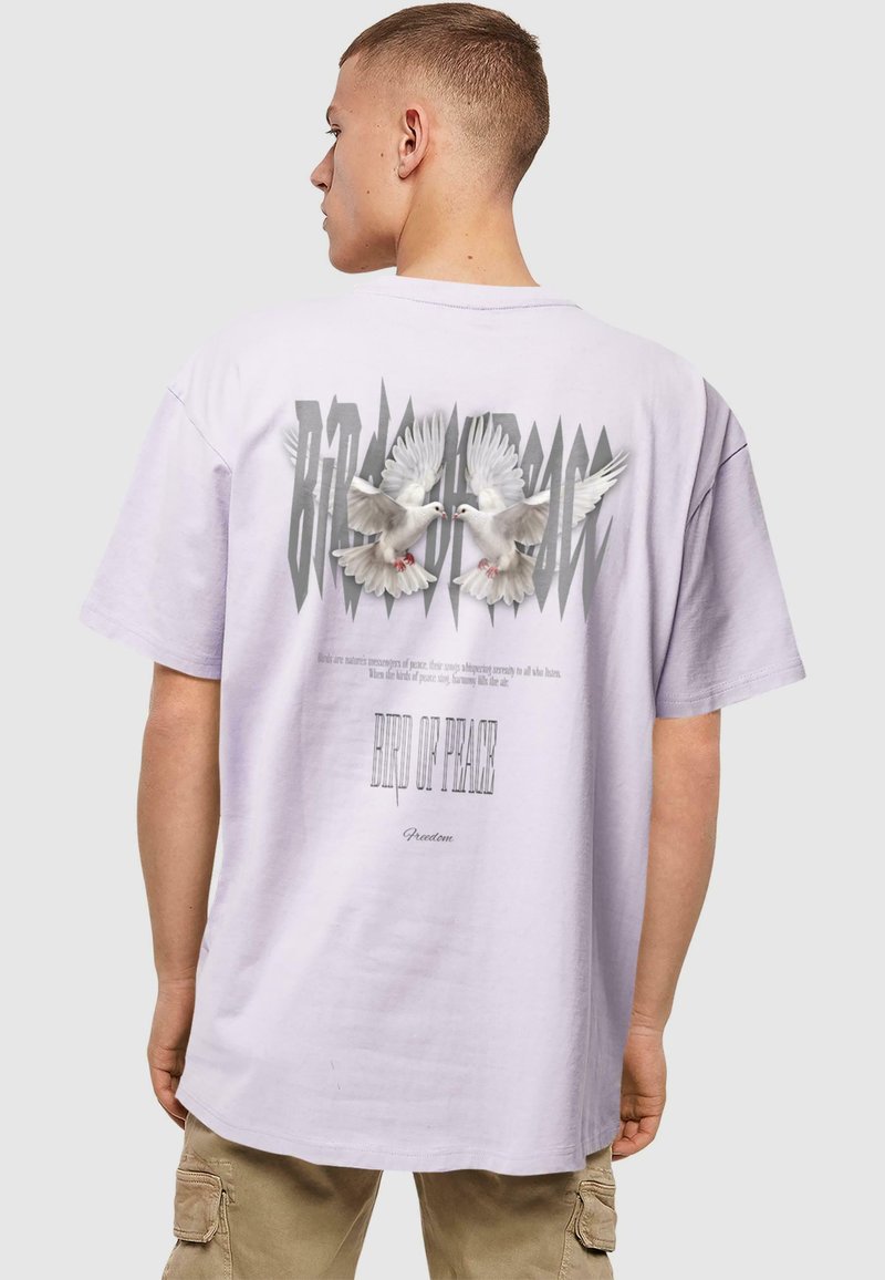 Lilla oversized t-shirt med et grafik af to hvide duer og stiliseret tekst "FUGL AF FRED" på bagsiden. Korte ærmer, lavet af bomuldsmateriale.