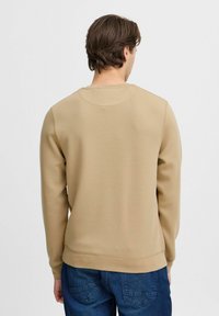 Beige sweatshirt med rund halsudskæring, ribbede manchetter og kant. Glat tekstur med en afslappet pasform, båret over blå denimbukser.